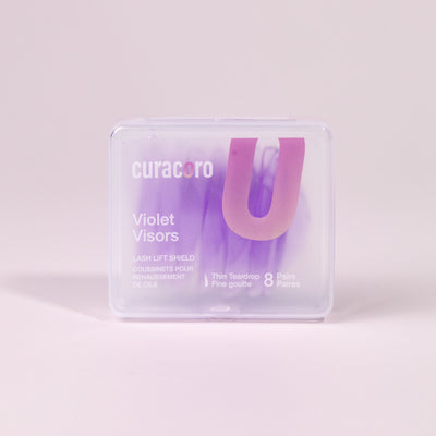 Curacoro Violet Visors lash lift shields in a transparent case featuring 8 pairs of thin teardrop-shaped shields. on a soft pink background. 