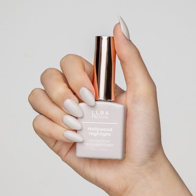 Hand holding a bottle of LLBA Pronail Hollywood Highlight Nail Gel Polish, featuring a soft white shade, with matching nail color on the hand, against a white background.