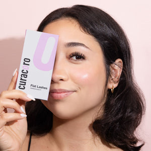 A woman with a radiant complexion holds a Curacoro Flat Lashes product box near her face, with a soft pink background.