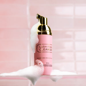 Lash Foam Cleanser bottle surrounded by white foam, set against a glossy pink tiled background.