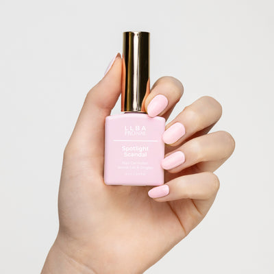 Hand holding a bottle of LLBA ProNail Spotlight Scandal nail gel polish, with nails painted in a matching soft pink shade. The bottle has a gold cap and the product name is clearly visible on the front.