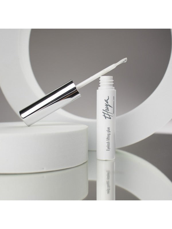 Open bottle of eyelash lifting glue with a shiny silver cap and white wand, placed on reflective white circular props.