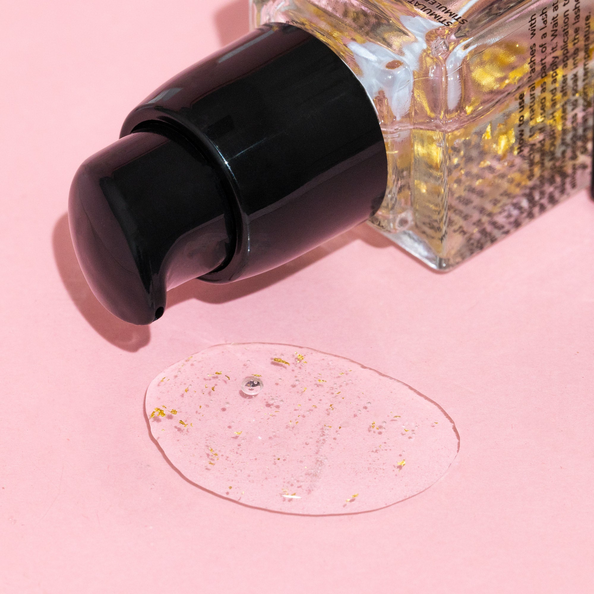 A close-up of a pump bottle with gold flakes, alongside a drop of clear gel with gold specks on a pink background.