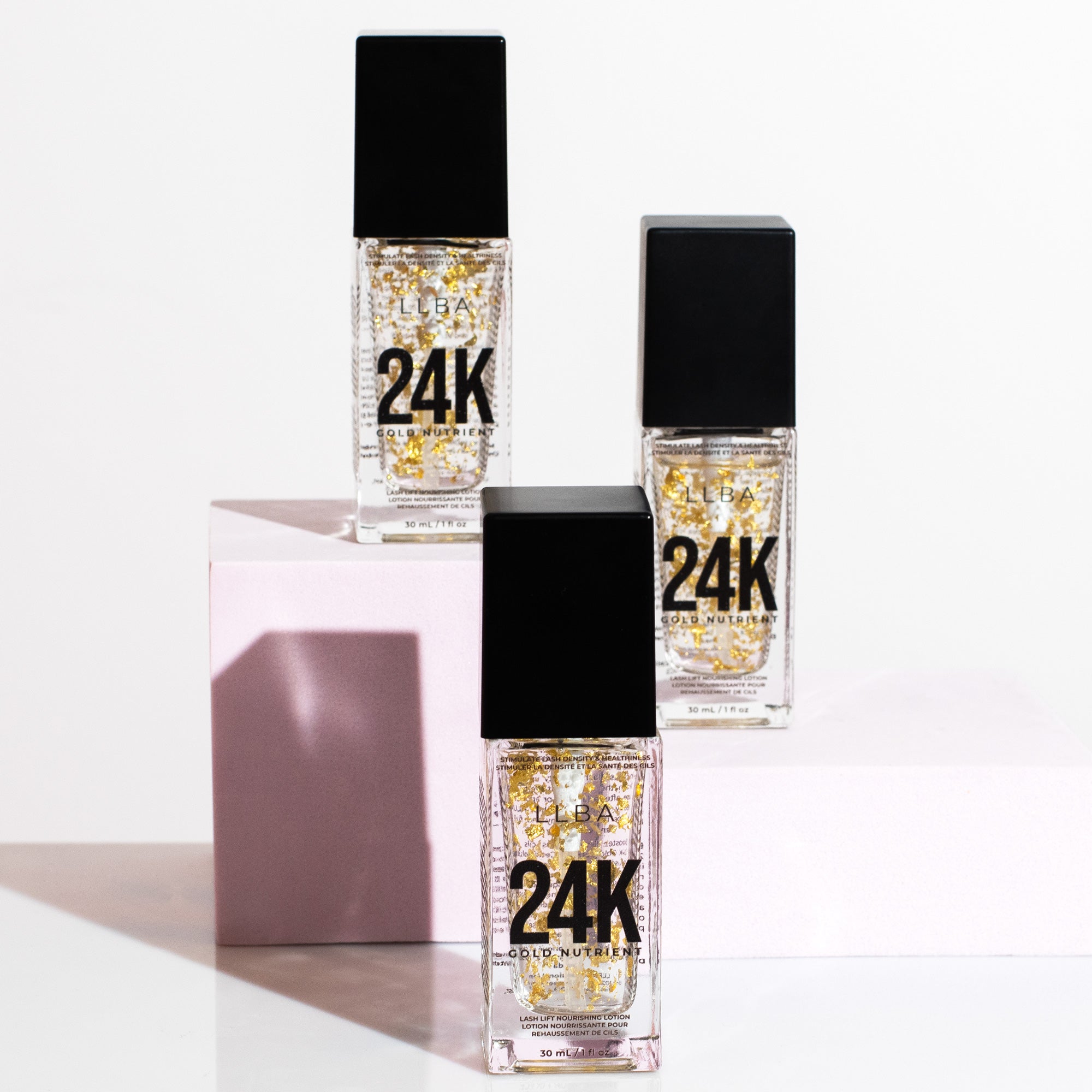 Three bottles with gold flakes inside, arranged on pink and white platforms, casting shadows on a bright background.