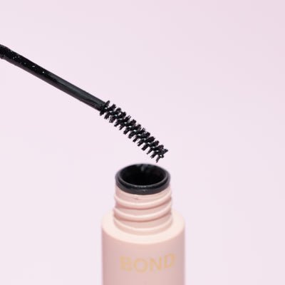 Close-up of the 'Bond' applicator wand for 'Quickie by LLBA' lash adhesive, showing a black, bristled brush above the open container on a light pink background.